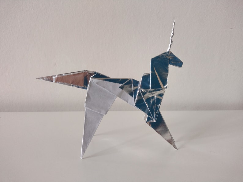 May include: A silver origami unicorn with a curled horn. The unicorn is standing on a white surface.
