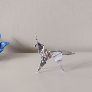 May include: A silver origami unicorn stands on a white surface. The unicorn has a long, pointed horn and a flowing mane.