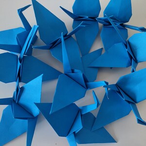 May include: A group of blue origami cranes, folded from paper, arranged in a circular pattern on a white surface.