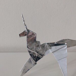 May include: A silver origami unicorn with a curled horn. The unicorn is made of folded paper and has a white underside.
