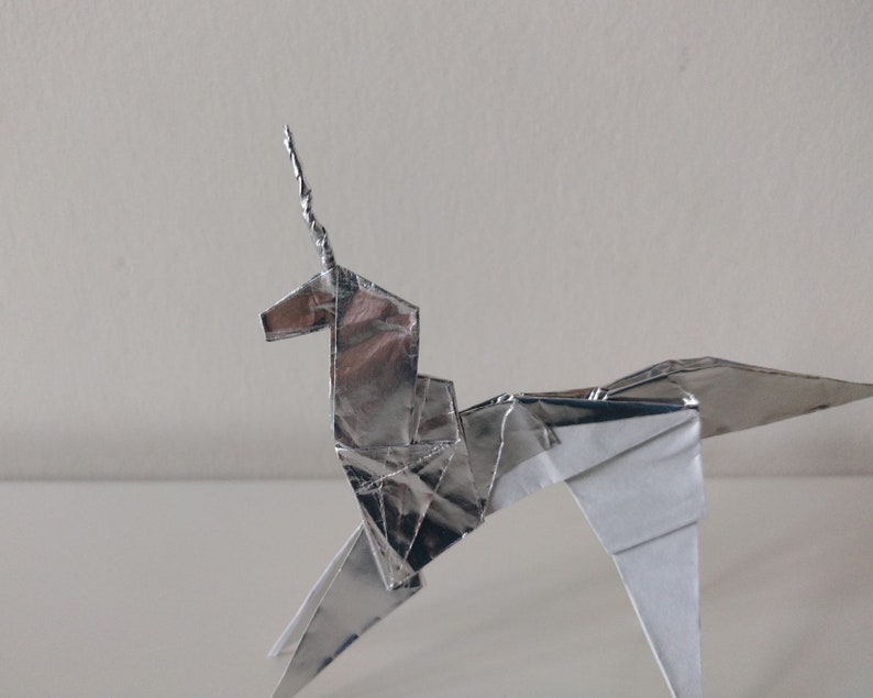 May include: A silver origami unicorn with a curled horn. The unicorn is made of folded paper and has a white underside.