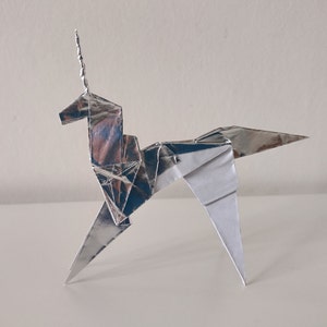 May include: A silver origami unicorn with a curled horn. The unicorn is standing on a white surface.