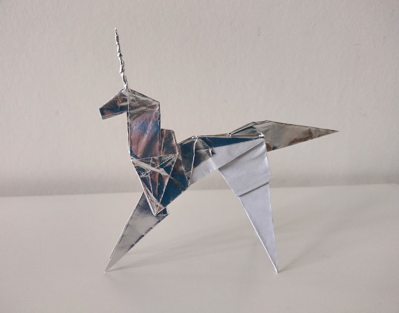 May include: A silver origami unicorn with a curled horn. The unicorn is standing on a white surface.
