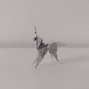 May include: A silver origami unicorn, folded from a single sheet of paper, stands on a white surface. The unicorn has a long, pointed horn and a flowing mane.