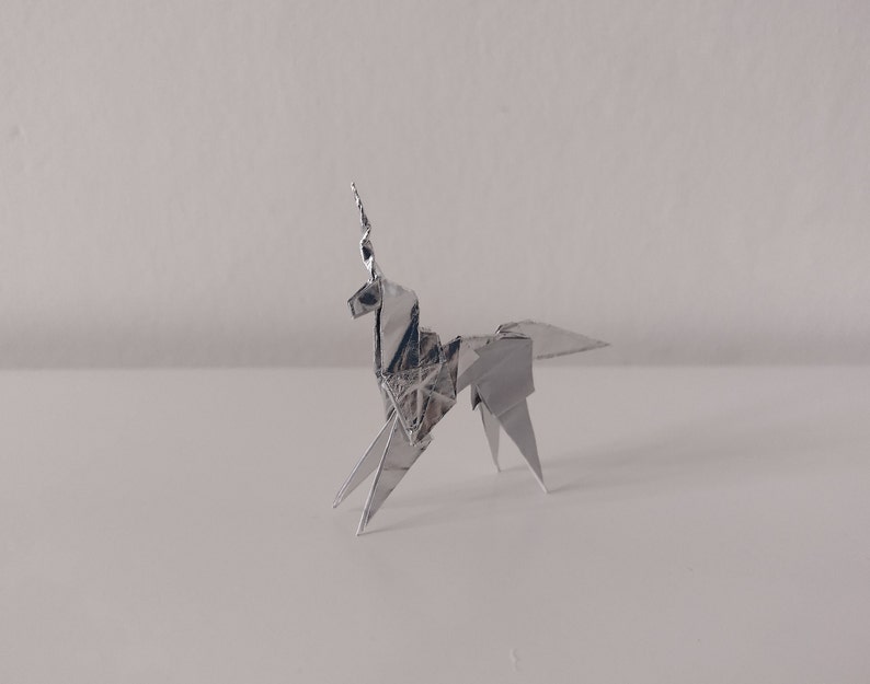 May include: A silver origami unicorn, folded from a single sheet of paper, stands on a white surface. The unicorn has a long, pointed horn and a flowing mane.