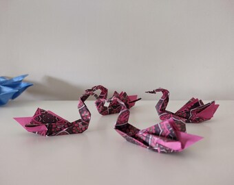 May include: Origami swans in shades of pink and burgundy with a patterned design. The paper swans are arranged on a white surface. A blue origami flower is in the background.