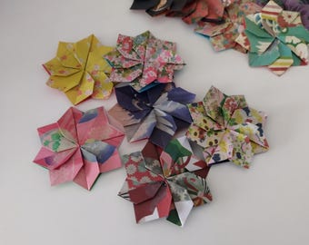 May include: A collection of colorful origami flowers in various sizes and patterns. The flowers are made from patterned paper in shades of pink, yellow, blue, and green. The origami flowers are arranged on a white surface.