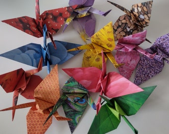 May include: A collection of ten origami paper cranes, each folded from different patterned paper. The cranes are arranged in a circle on a white surface. The patterns include flowers, pebbles, and a peacock design.