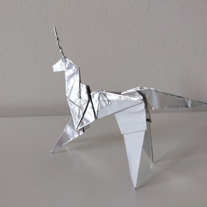 May include: A silver origami unicorn with a long, pointed horn. The unicorn is standing on a white surface.