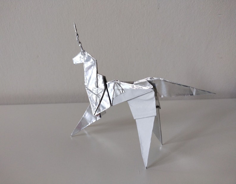 May include: A silver origami unicorn with a long, pointed horn. The unicorn is standing on a white surface.