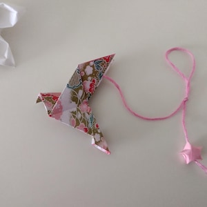 May include: Origami bird with pink string and star-shaped charm. The bird is made of patterned paper with pink, gold, and floral designs. A white paper flower is in the background. The origami bird is a decorative item.