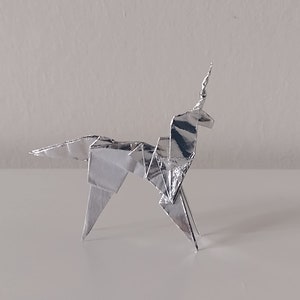 May include: A silver origami unicorn, folded from a single sheet of paper, stands on a white surface.