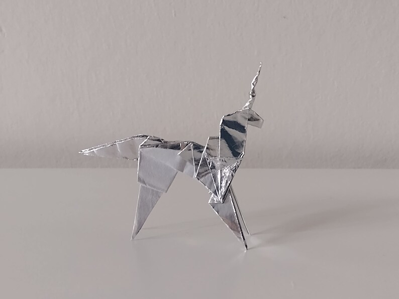May include: A silver origami unicorn, folded from a single sheet of paper, stands on a white surface.