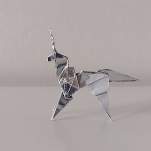 May include: A silver origami unicorn, folded from a single sheet of paper, stands on a white surface. The unicorn's horn is pointed and its mane is folded into a series of small triangles.