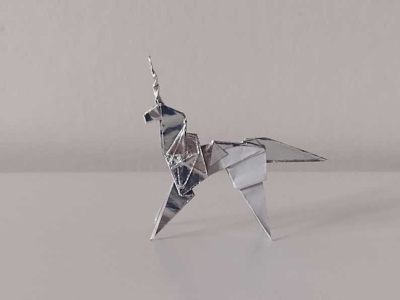May include: A silver origami unicorn, folded from a single sheet of paper, stands on a white surface. The unicorn's horn is pointed and its mane is folded into a series of small triangles.