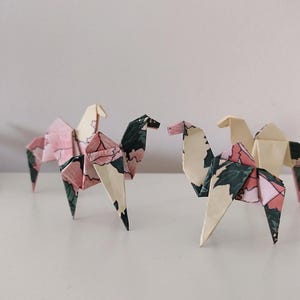 Peut inclure: Trois figurines de chameaux en origami, aux motifs floraux roses, verts et crème. Les sculptures en papier sont posées sur une surface blanche, mettant en valeur les plis et les motifs complexes. Les chameaux se font face.