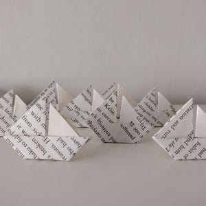 Op de afbeelding: Een verzameling kleine origamibootjes gevouwen van witte boekpagina's met zwarte gedrukte tekst. De papieren vaartuigen hebben puntige zeilen en rompen, gerangschikt in een groep op een plat oppervlak, met een blauwe vorm aan de linkerkant.