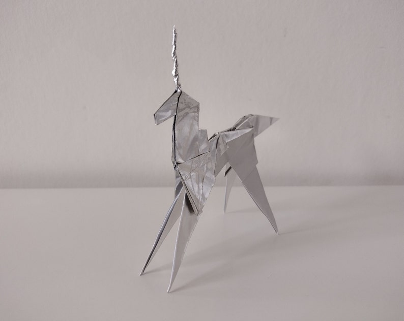 May include: A silver origami unicorn sculpture. The unicorn is standing on a white surface.