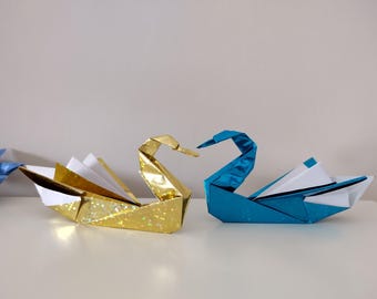 May include: Two origami swans, one gold and one teal, are displayed on a white surface. The gold swan has a shimmering finish, while the teal swan has a glossy appearance. Both swans are folded with intricate detail.