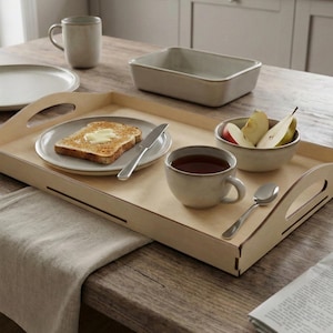 May include: A wooden serving tray with handles holds a plate of toast with butter, a cup of tea, a bowl of fruit, and a spoon. A mug, a plate, and a rectangular dish are also on the wooden table. The items are neutral in color.