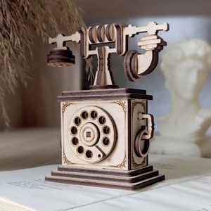 May include: A wooden model of a vintage rotary phone with a brown base and a detailed dial. The phone has a wooden receiver and a crank on the side.