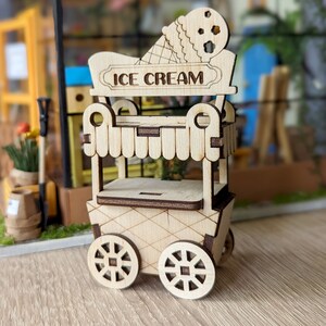 Op de afbeelding: Een houten ijskar met wielen, een bord met de tekst "Ice Cream" en een decoratieve ijshoorn bovenop.