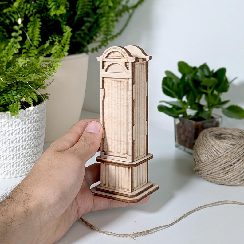 May include: A wooden model of a grandfather clock with a detailed design. The clock is made of light brown wood and has a white face. The clock is about 10 inches tall.