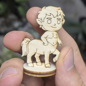 Könnte beinhalten: Eine Holzfigur eines Zentauren mit Cartoon-Design. Der Zentaur hat einen menschlichen Oberkörper und einen Pferdekörper. Die Figur ist hellbraun und steht auf einem kleinen, runden Sockel. Der Zentaur wird in der Hand einer Person gehalten.