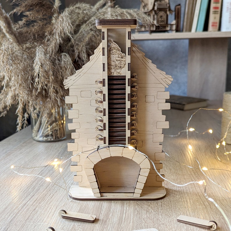 May include: A wooden advent calendar shaped like a house with a chimney and a fireplace. The numbers 11 through 24 are visible on the side of the house. A small figure of Santa Claus is visible in the chimney.