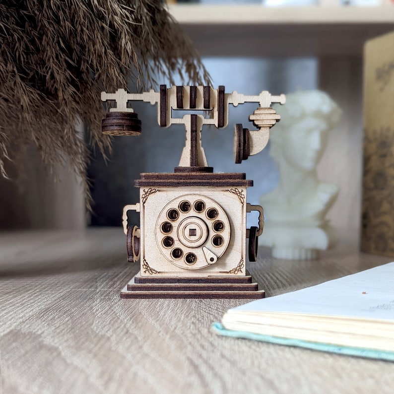 Op de afbeelding: Een houten model van een vintage draaitelefoon met een bruine basis en een cr&egrave;mekleurige draaiknop. De telefoon heeft een draaiknop aan de zijkant en een hoorn aan de bovenkant.
