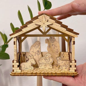 Puede incluir: Escena de la Natividad de madera con un establo con forma de estrella con figuras talladas de María, José, el niño Jesús, un burro y una oveja. La madera de color marrón claro tiene un acabado natural, creando un tema rústico y religioso.