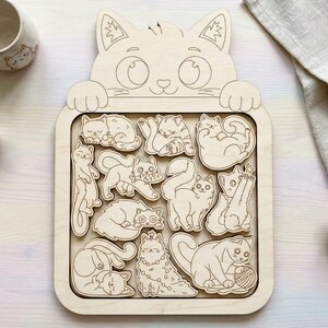 Op de afbeelding: Houten kattenpuzzel met 15 individuele kattenstukken. De puzzel toont verschillende kattenposities, waaronder slapen, spelen en staan. Het frame heeft de vorm van een kattenkop met grote ogen en poten. De puzzel is gemaakt van lichtgekleurd hout.