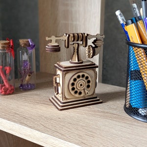 Op de afbeelding: Een houten model van een vintage draaitelefoon met een gedetailleerd ontwerp. De telefoon heeft een ronde draaiknop met cijfers en een hoorn aan de zijkant. Het model is geverfd in een natuurlijke houtkleur.