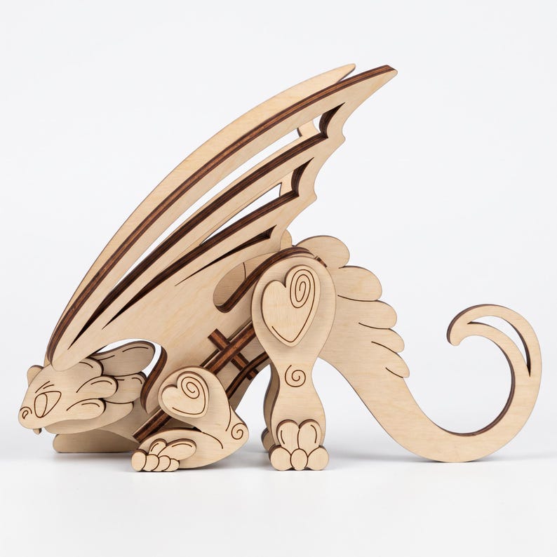 Side profile view of 3D laser cut plywood crouching dragon model with pointed wings and curled tail on white background.
