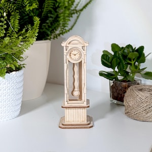 May include: A wooden model of a grandfather clock with a white face and black hands. The clock is standing on a white surface.