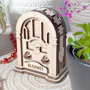 Könnte beinhalten: Ein Holzmodell eines Vintage-Radios mit hellbraunem Finish und dunkelbraunen Akzenten. Das Wort "RADIO" ist auf der Vorderseite aufgedruckt. Das Radio hat zwei Knöpfe und ein Zifferblatt. Das Design erinnert an Radios aus dem frühen 20. Jahrhundert.
