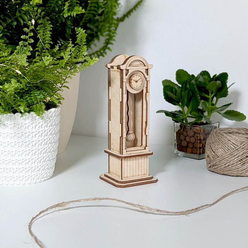 May include: A wooden model of a grandfather clock with a pendulum and a clock face. The clock is made of light brown wood and has a white background.