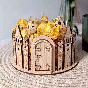 Puede incluir: Un recipiente de madera para dulces con forma de castillo con una puerta y una valla. El recipiente está lleno de caramelos amarillos y blancos.