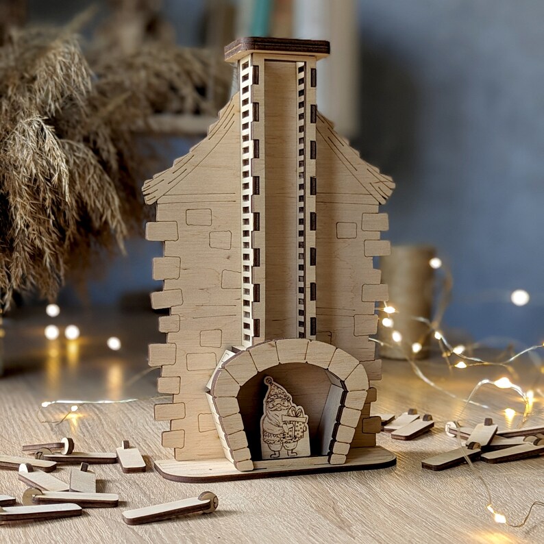 May include: A wooden Christmas house with a chimney and a fireplace. The house is decorated with a Santa Claus figure holding a gift. The house is made of laser-cut wood and is ready to be assembled.