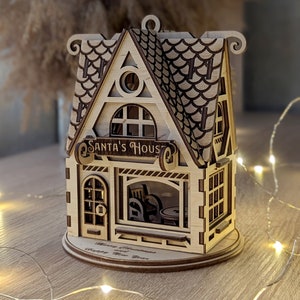 May include: A wooden Christmas house ornament with a sign that reads "Santa's House". The house has a window with a view of a fireplace and a Christmas tree. The ornament is on a wooden base with the words "Merry Christmas and Happy New Year".