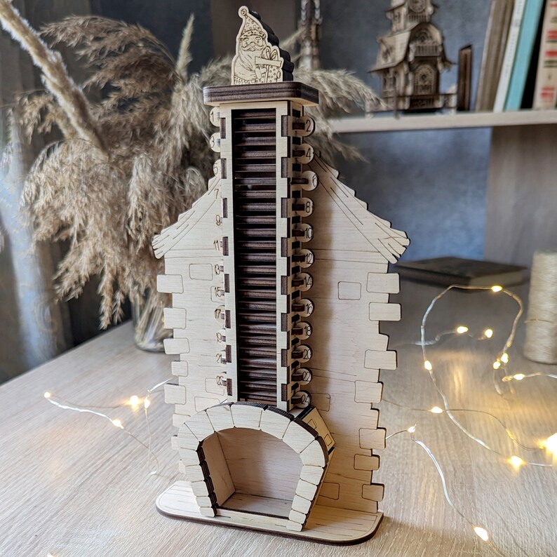 May include: A wooden Christmas countdown calendar shaped like a house with a chimney. The chimney has a sliding door with numbers 1 through 24 printed on it. A Santa Claus figure is on top of the chimney.