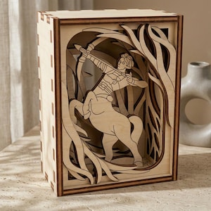Könnte beinhalten: Holzkiste mit einem geschichteten Design, das einen Bogenschützen-Zentaur zeigt. Das Kunstwerk ist aus hellem Holz gelasert, mit dunkleren Holzakzenten am Rahmen. Die Kiste ist rechteckig und steht aufrecht.