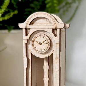 May include: A wooden grandfather clock model with a face showing the time. The clock is made of light brown wood and has a traditional design.
