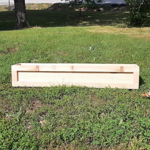 May include: A wooden planter box, made of natural wood, sits on a grassy lawn. The planter is rectangular and has a simple design.