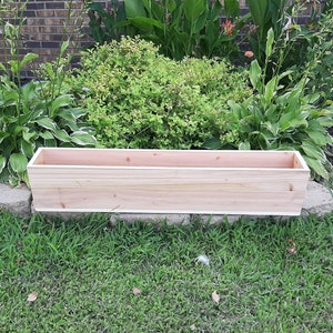 May include: A natural wood planter box, made of cedar, sits on a bed of green grass. The planter is rectangular and has a simple design.