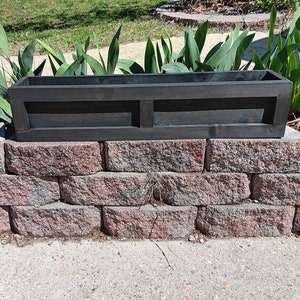 May include: A black wooden planter box with two compartments. The planter is sitting on a brick wall.