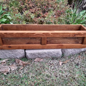 May include: A long, rectangular wooden planter box with a dark brown stain. The planter has a simple design with a raised lip around the top and a decorative panel in the center.