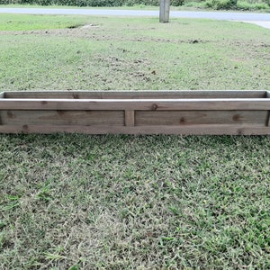 May include: A long, rectangular wooden planter box with a weathered gray finish. The planter has a simple design with a single panel on each side and a raised lip around the top.