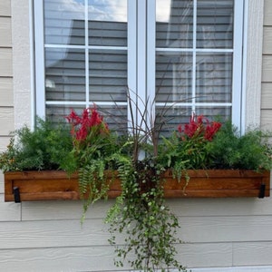 May include: A wooden window box filled with a variety of plants and flowers. The box is stained a dark brown and is mounted below a white window. Red flowers and green foliage are the primary colors.