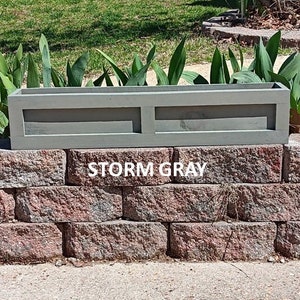 May include: A rectangular storm gray planter box with a recessed design, sitting atop a brick wall. The text "STORM GRAY" is visible on the wall. Green plants are in the background.